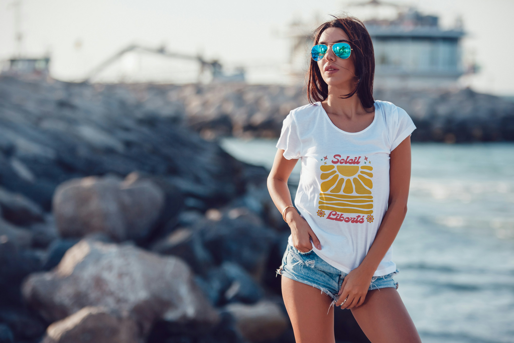 T-shirt blanc oversize Soleil & Liberté avec motif rétro groovy 70s jaune et orange, femme portant lunettes de soleil bleues .